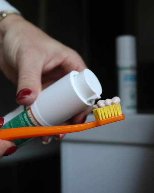 Tube de dentifrice blanc, bouchon ouvert et brosse à dents orange en premier plan.